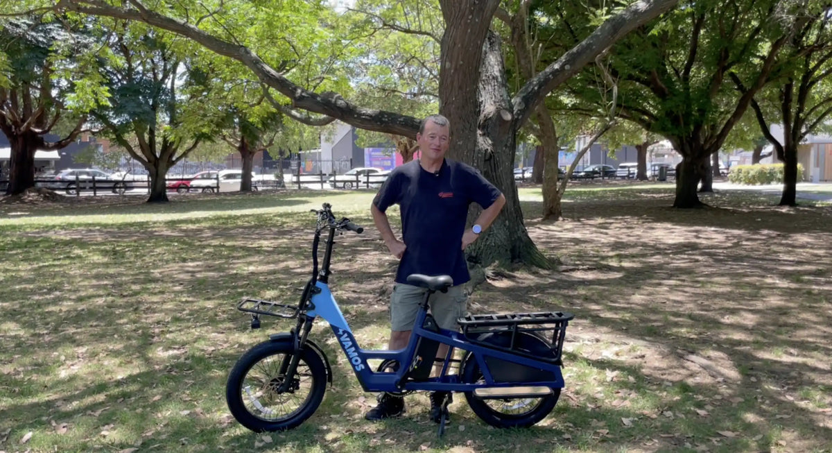 Man standing next to a blue Vamos longtail cargo e-bike with front rack and heavy-duty rear passenger rack.