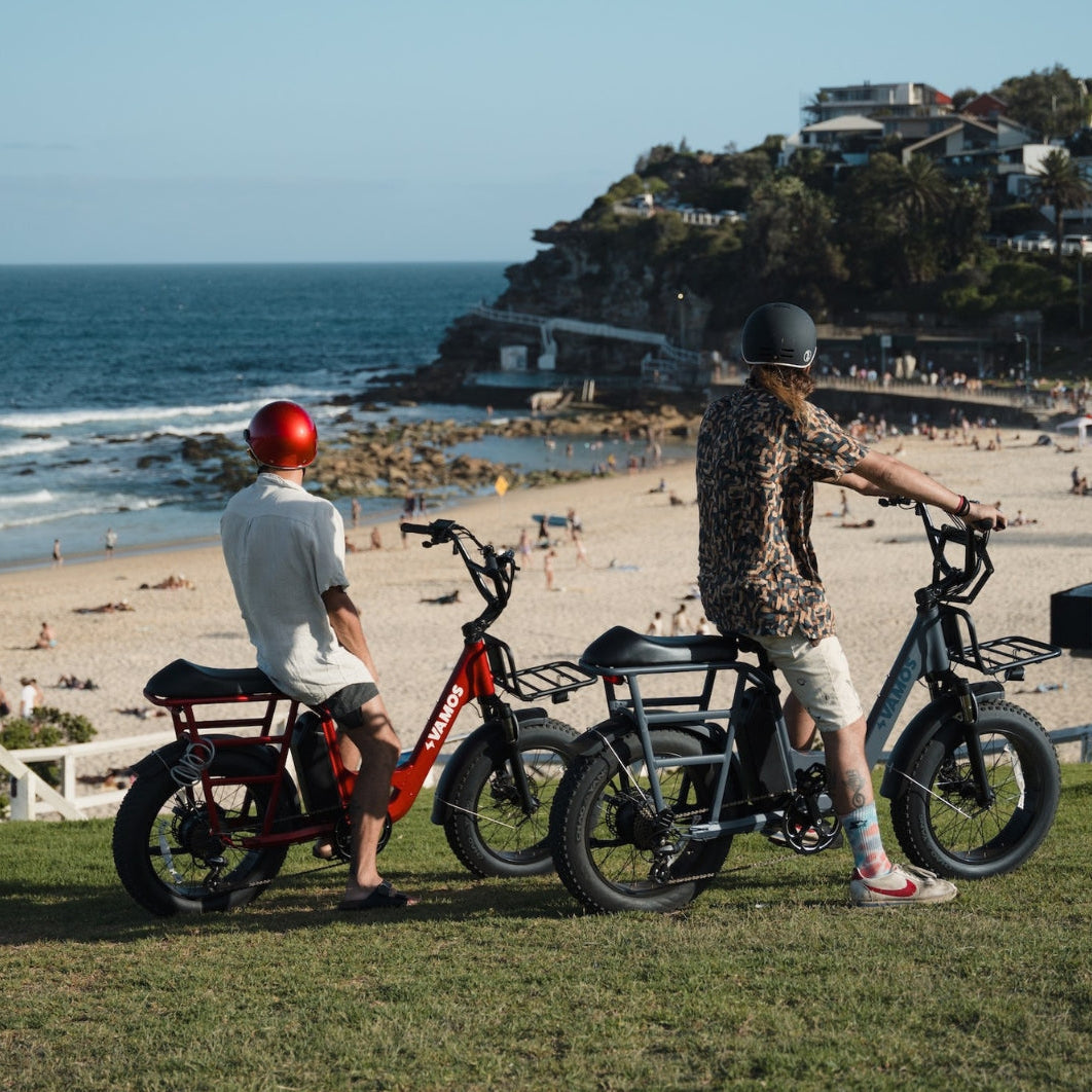 Two riders on Vamos electric bicycles overlooking a crowded beach