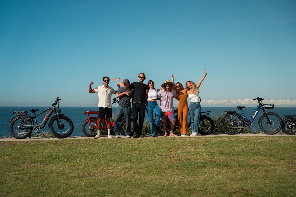 Group of friends posing with various Vamos electric bikes and e-trikes along a scenic coastal park headland.