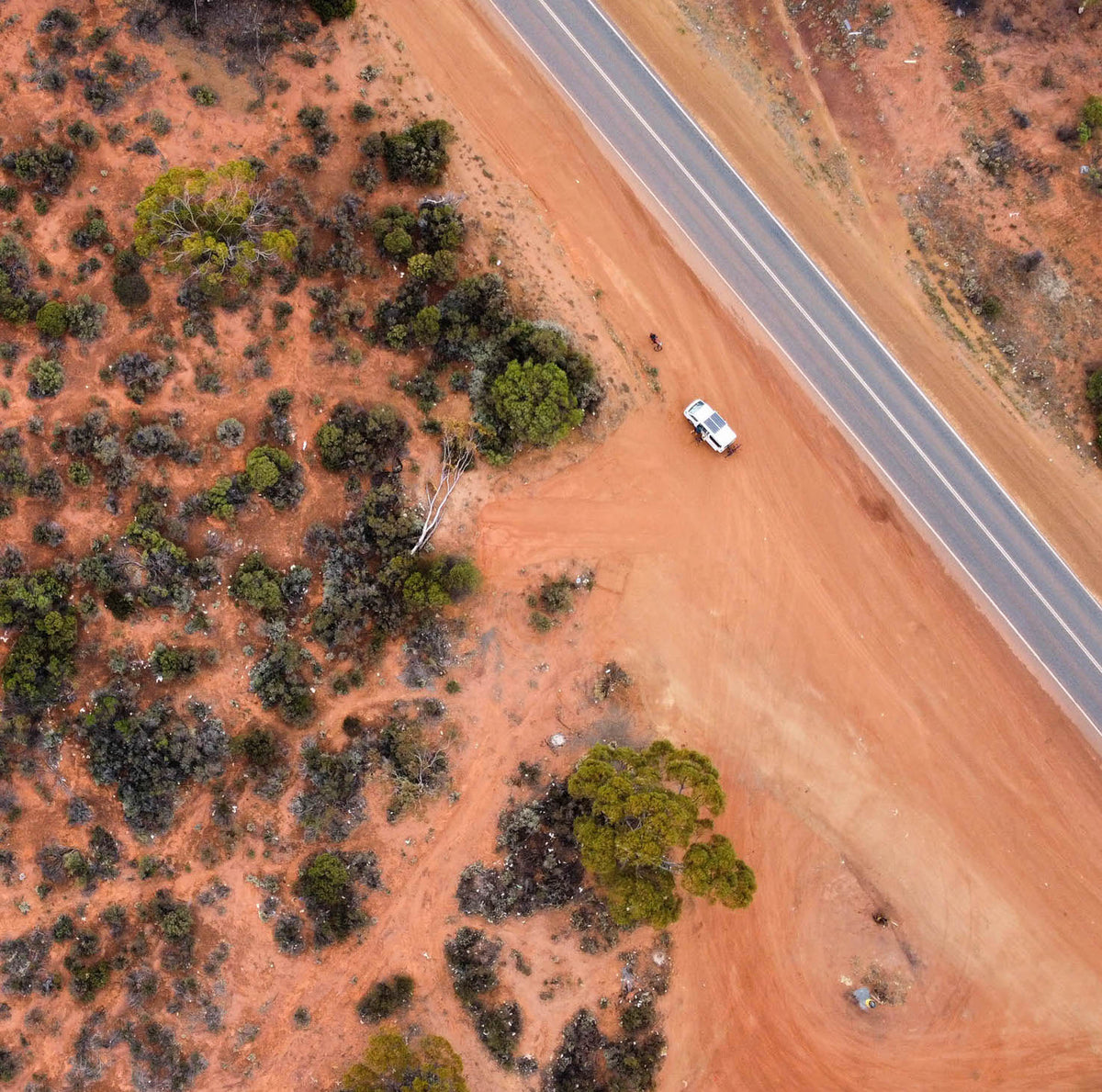An aerial view of a white SUV parked on a red dirt shoulder next to a paved highway in the Australian Outback.