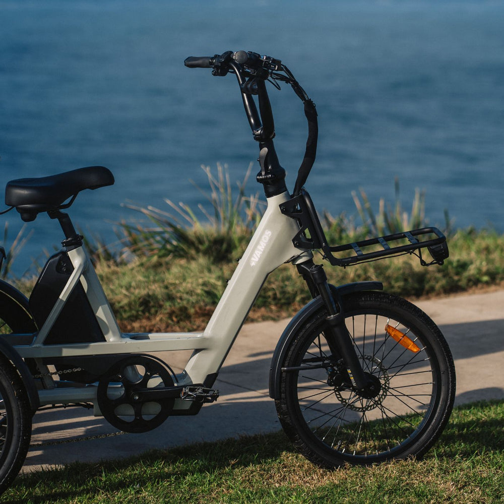 Side profile photo of a cream-colored Vamos electric cargo trike with front rack parked along a scenic oceanfront trail.