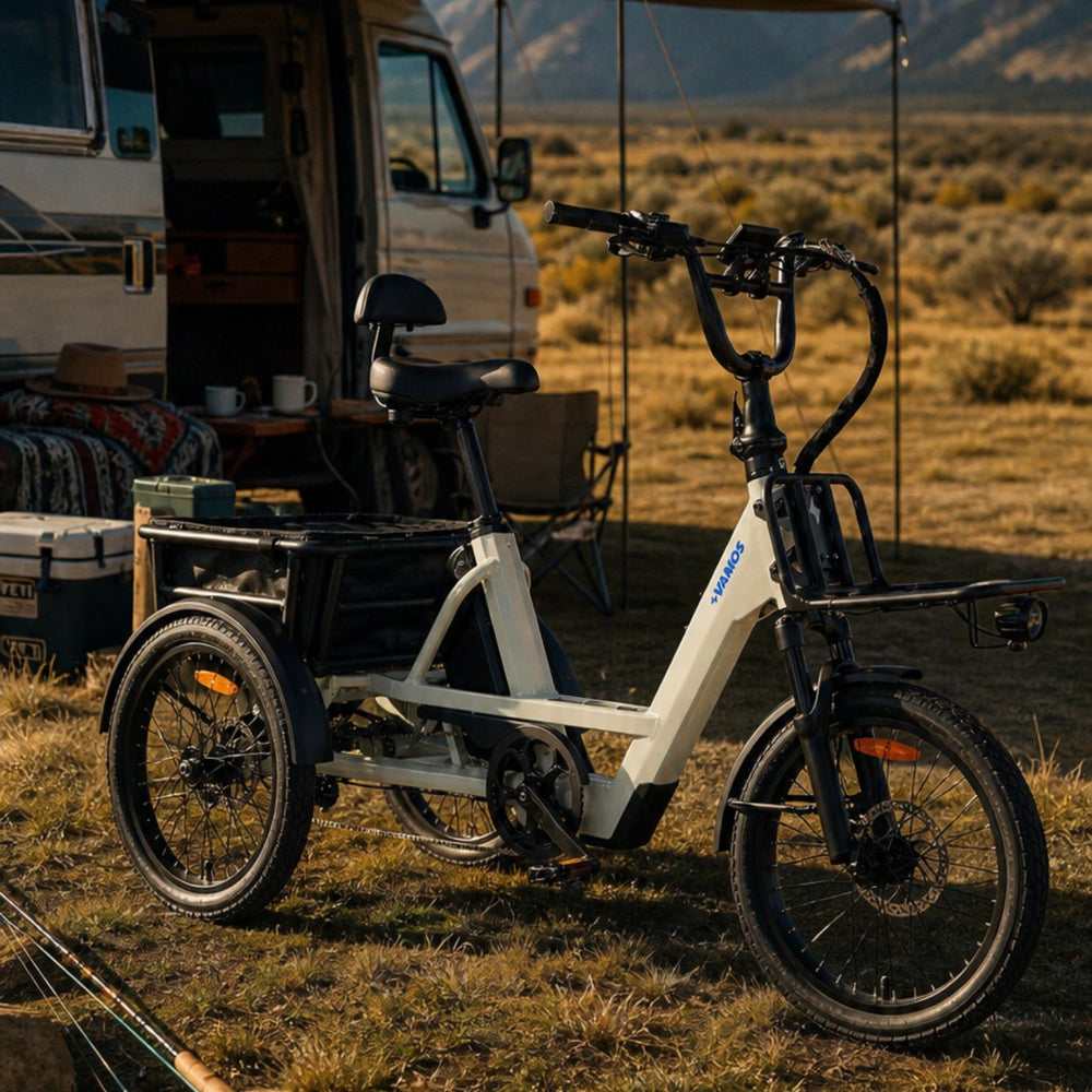 Outdoor lifestyle photograph of a cream-colored Vamos electric cargo tricycle parked at a camper van campsite with open fields in the background.