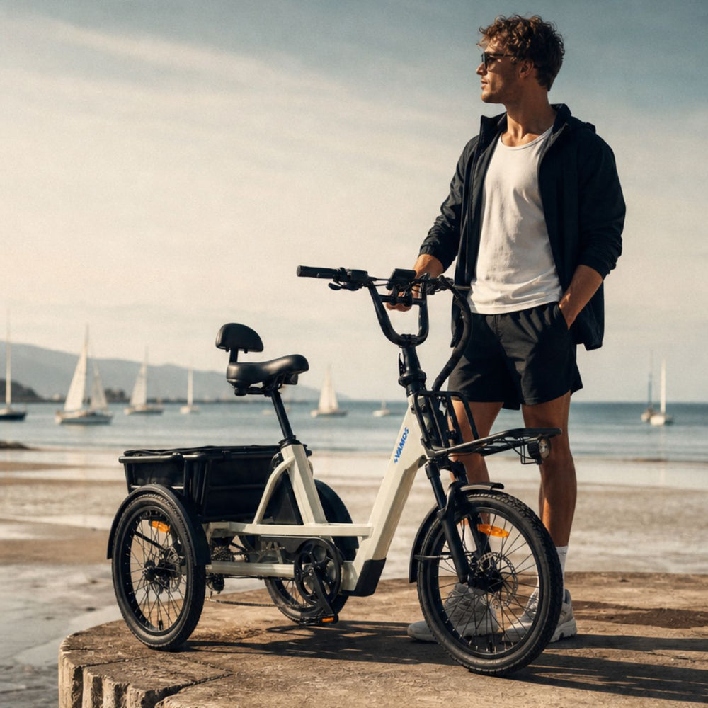 Outdoor portrait of a man standing beside a cream-colored Vamos electric cargo tricycle at a coastal beach overlook with sailboats in the background.