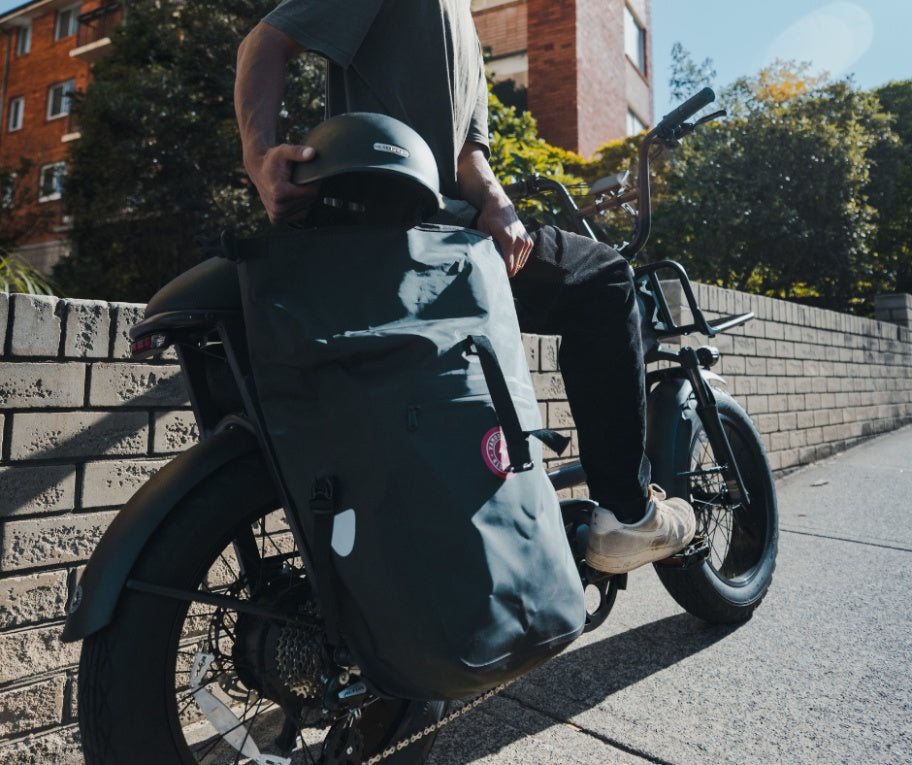A person sits on an electric cargo bike holding a helmet, featuring a large Vamos Waterproof Pannier Bag for Bike