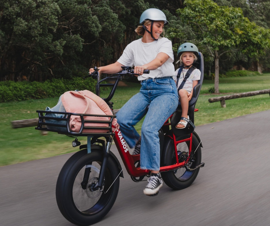 A woman rides a red Vamos electric cargo bike with a child riding safely behind her in a grey Thule Yepp 2 Maxi Child Bike Seat.