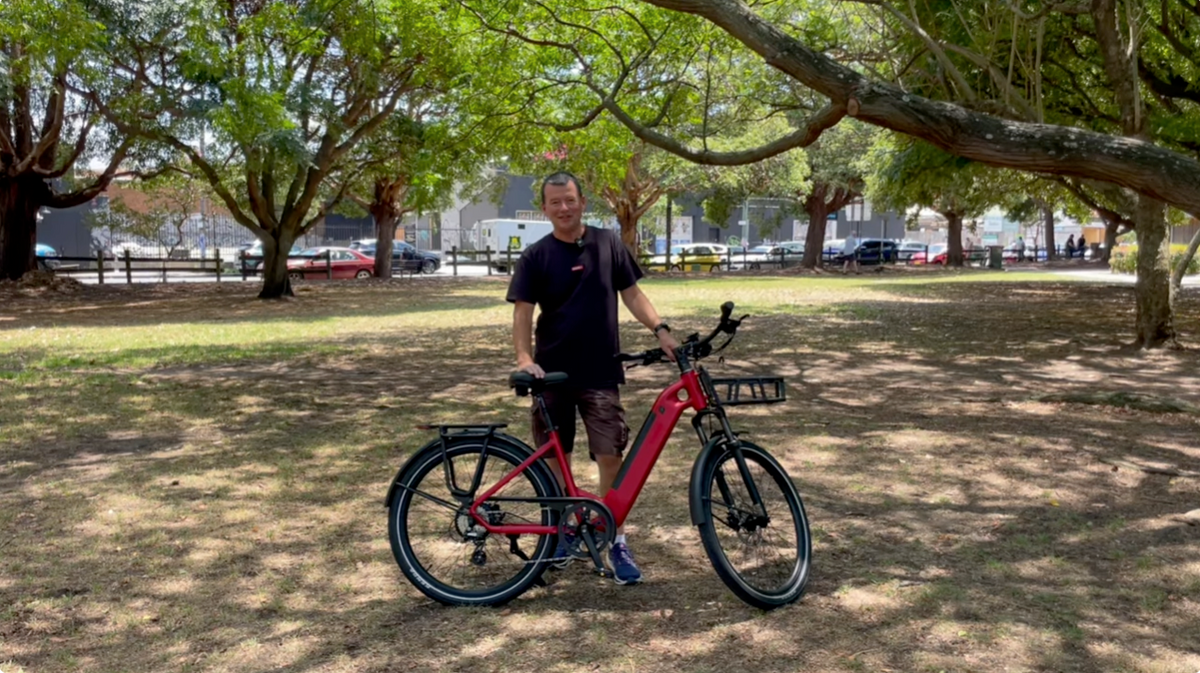 Man standing with a red Vamos electric bike with front and rear racks in a grassy park setting.