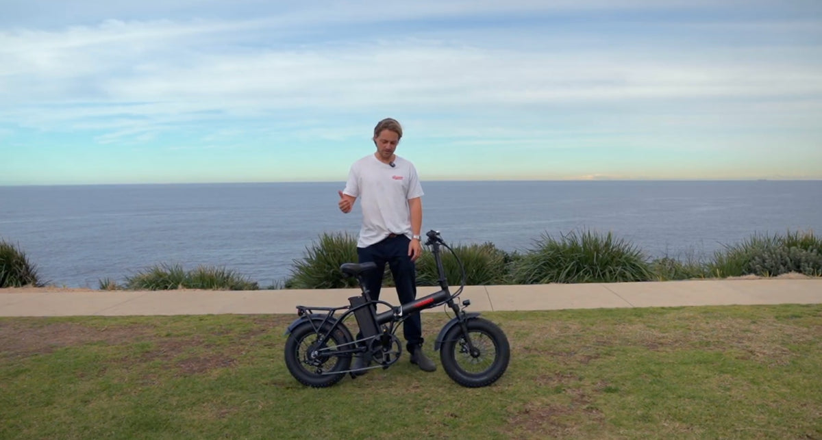 A person standing next to a compact, Vamos Doblez Foldable E-Bike by the coast.