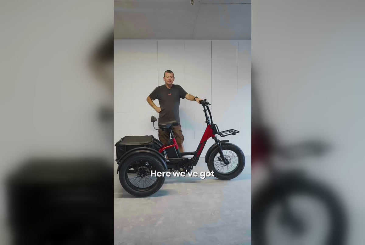 Man standing beside a red and black Vamos electric tricycle in front of a white wall.
