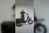 Man standing beside a red and black Vamos electric tricycle in front of a white wall.