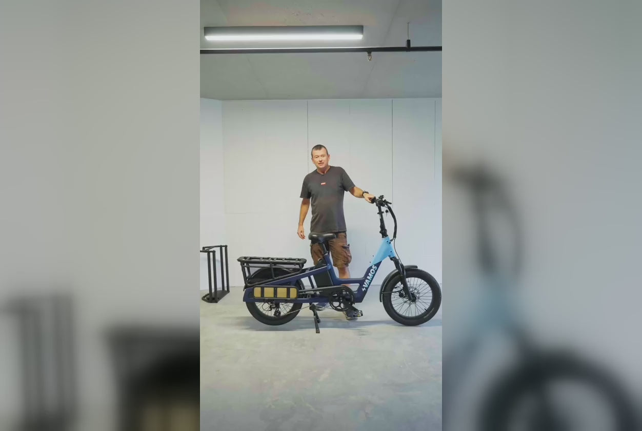 Man presenting a blue Vamos electric utility bike equipped with a rear cargo rack in a studio setting.