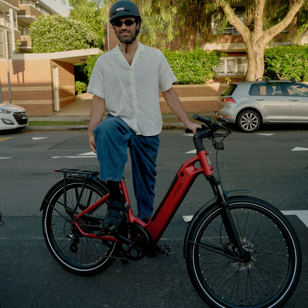 Man posing with a red Vamos step-through electric commuter bike equipped with a rear rack on a city street.