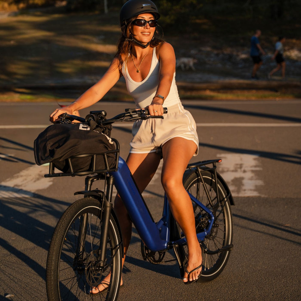 Woman riding a blue Vamos step-through electric commuter bike with a front basket at sunset.