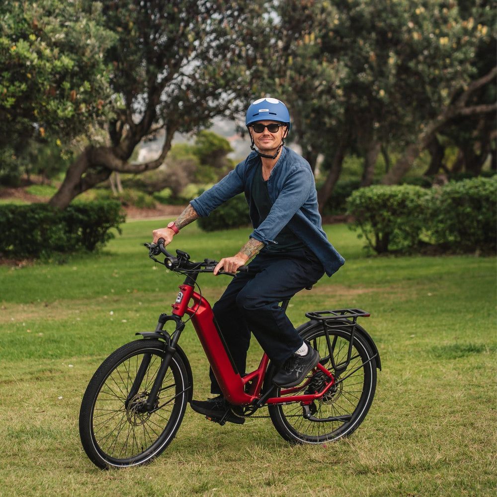 Man riding a red step-through Vamos electric bike across a grassy park path.