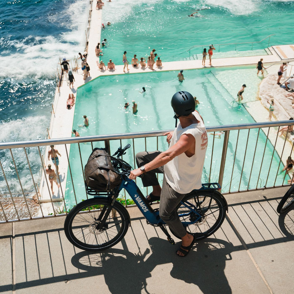 High-angle shot of rider on a blue Vamos e-bike with front cargo bag overlooking a coastal ocean pool.