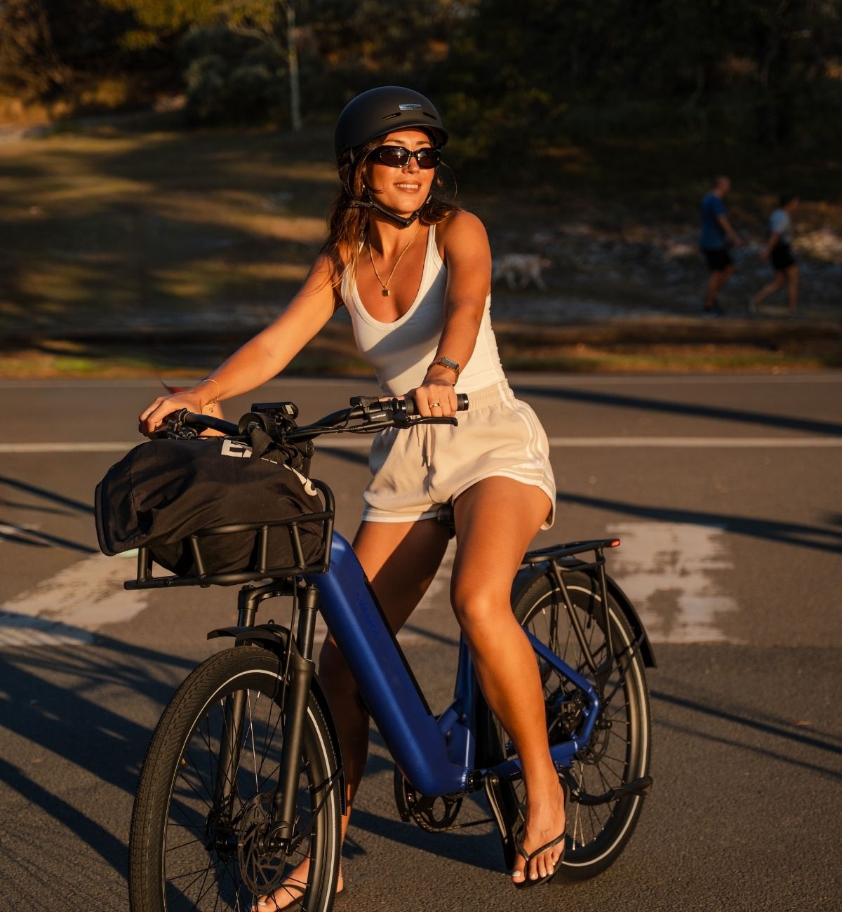 Front-three-quarter angle of rider on a blue Vamos step-through electric commuter bike with front basket and rear rack during golden hour.