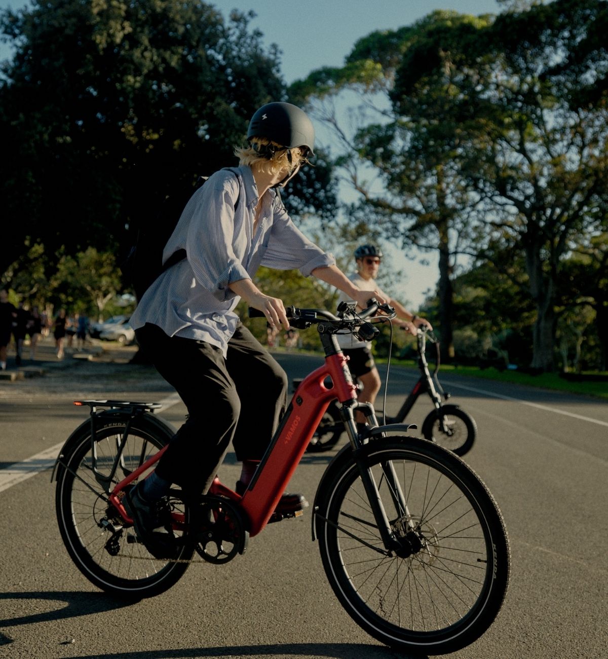 Low-angle side view of a cyclist riding a red step-through Vamos electric commuter bike through a tree-lined park.