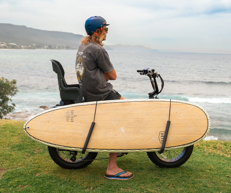 Vamos electric bicycle equipped with a side-mounted rack carrying a wooden surfboard along the coast.