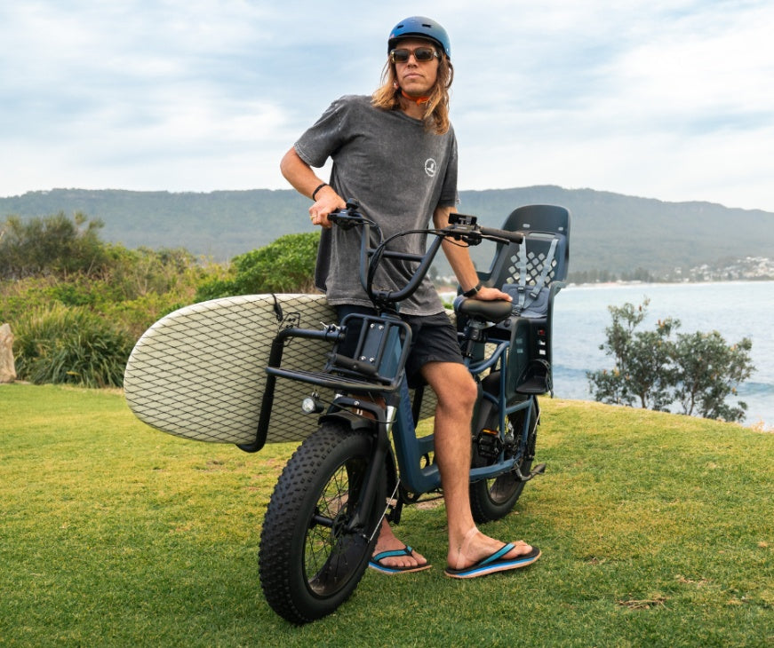 A person stands with a utility e-bike equipped with a front Surfboard Rack and a rear Thule Yepp 2 Maxi child seat.