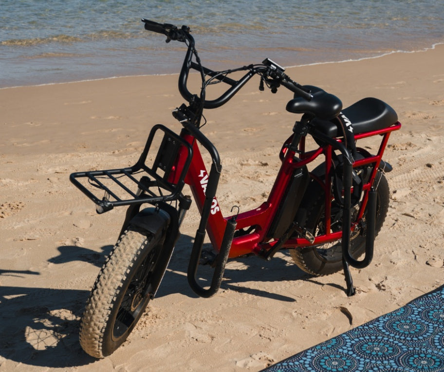 A red Vamos El Amigo compact cargo e-bike equipped with fat tires, a front cargo rack, and an attached side surfboard rack.