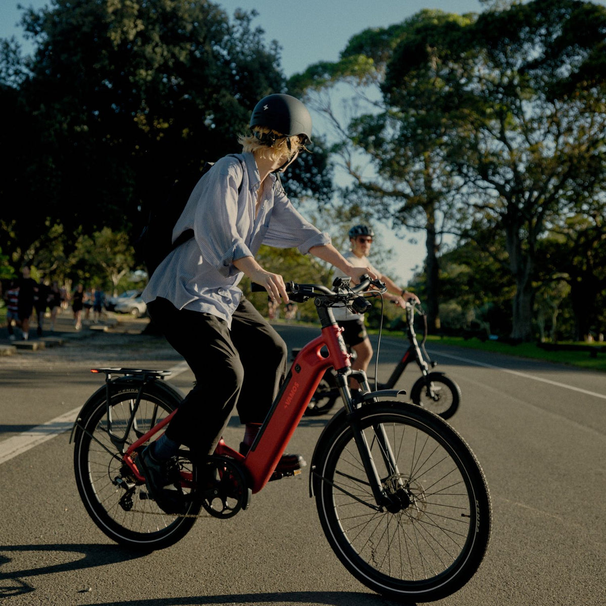 Side angle of a cyclist on a red Vamos step-through electric commuter bike with a rear rack riding in a park.