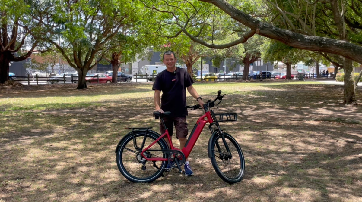 A man stands under a tree with a red Rapido City Daily commuter Ebike in a park